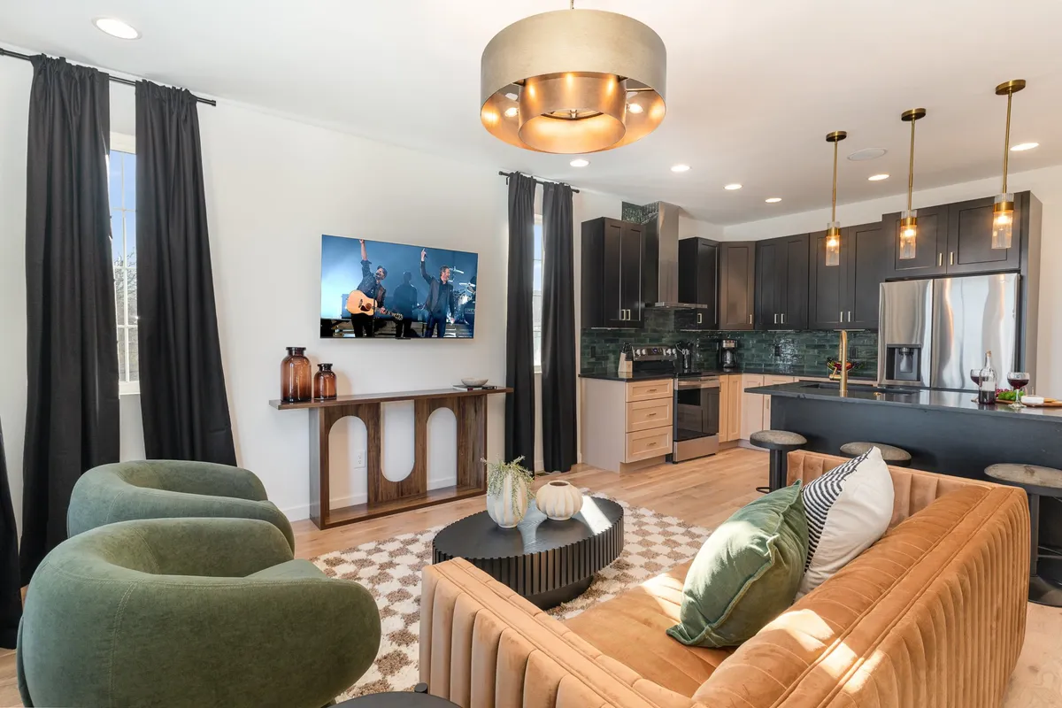 This image presents a spacious, open-plan living area with a stylish sofa accented with cushions, green accent chairs, and a television showing a live performance. The background reveals an adjacent dining area, capturing the seamless flow between living and eating spaces. Large windows let in natural light, enhancing the comfortable and lively mood.