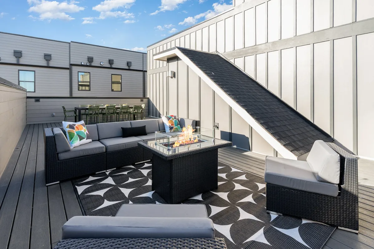 The image features a modern rooftop patio outfitted with comfortable outdoor seating arranged around a stylish fire pit atop a decorative rug. The warm glow of the fire and sunset lighting invite guests to relax or gather for evening conversations, all while taking in panoramic views of Nashville.