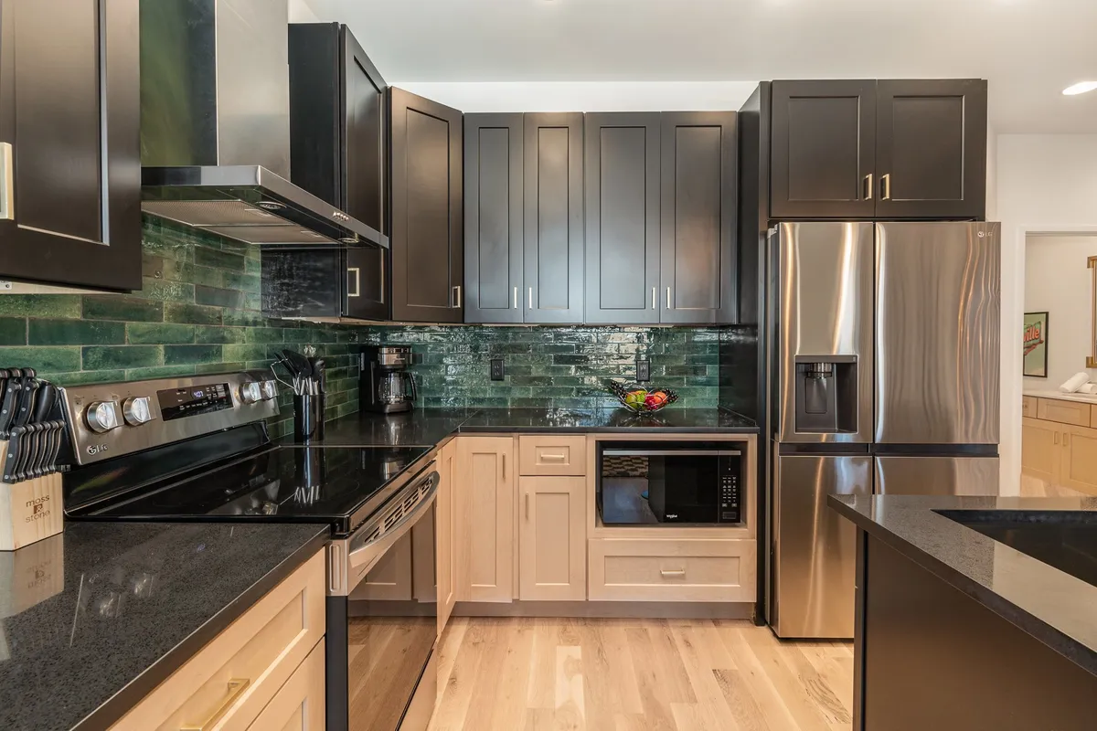 A stylish and functional kitchen with stainless steel appliances, green tile backsplash, a black countertop, and light wood cabinetry. The layout is designed for both casual family gatherings and meal preparation, featuring modern amenities.