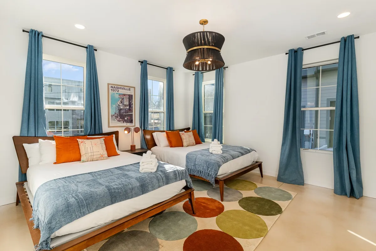 A comfortable bedroom featuring two beds with vibrant bedding, a colorful area rug, large windows that let in abundant natural light, a decorative light fixture, and framed wall art. The arrangement invites group stays and offers flexibility for different sleeping arrangements.