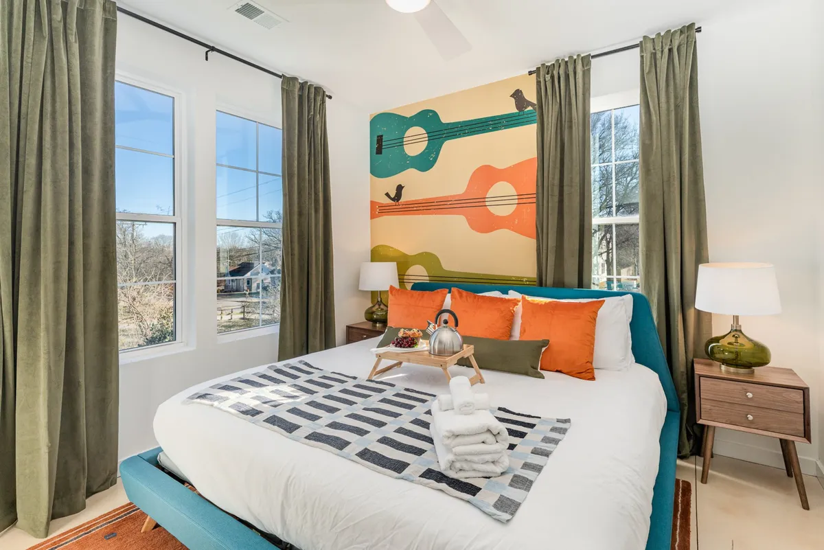 The room features a cozy bed setup with bright, colorful pillows, a large window letting in natural sunlight, and a striking guitar mural. The space feels playful yet restful, blending comfort with a creative vibe—ideal for relaxation and privacy.