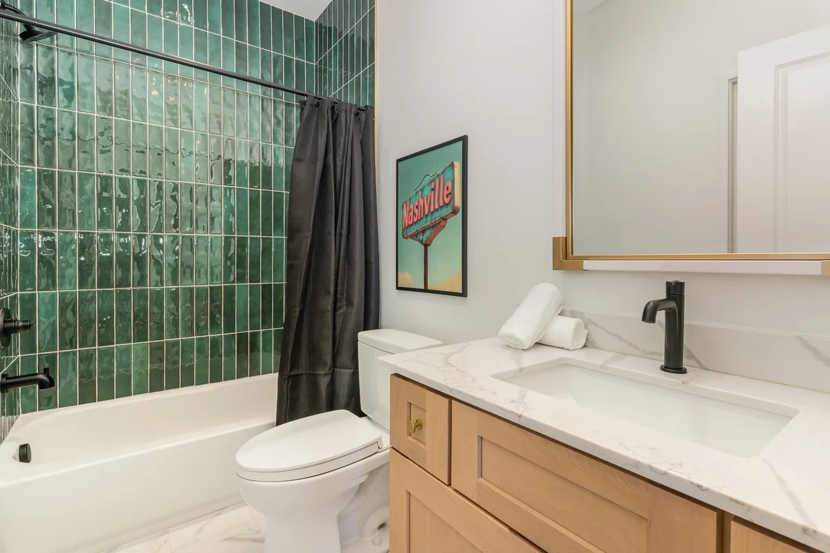 A contemporary bathroom with a green tiled wall, white countertop, black fixtures, and a piece of Nashville-themed artwork. The space is designed to offer comfort and style for guests.