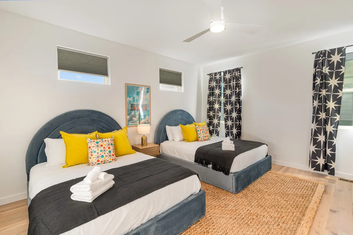 This image showcases two beds with colorful pillows, abstract artwork on the wall, and star-patterned curtains. The scene suggests a cheerful, relaxed atmosphere perfect for families or friends sharing a room. The large beds and soft lighting emphasize comfort and flexibility in sleeping arrangements.